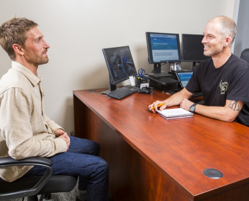 Man in beige shirt speaks with man in black shirt in office